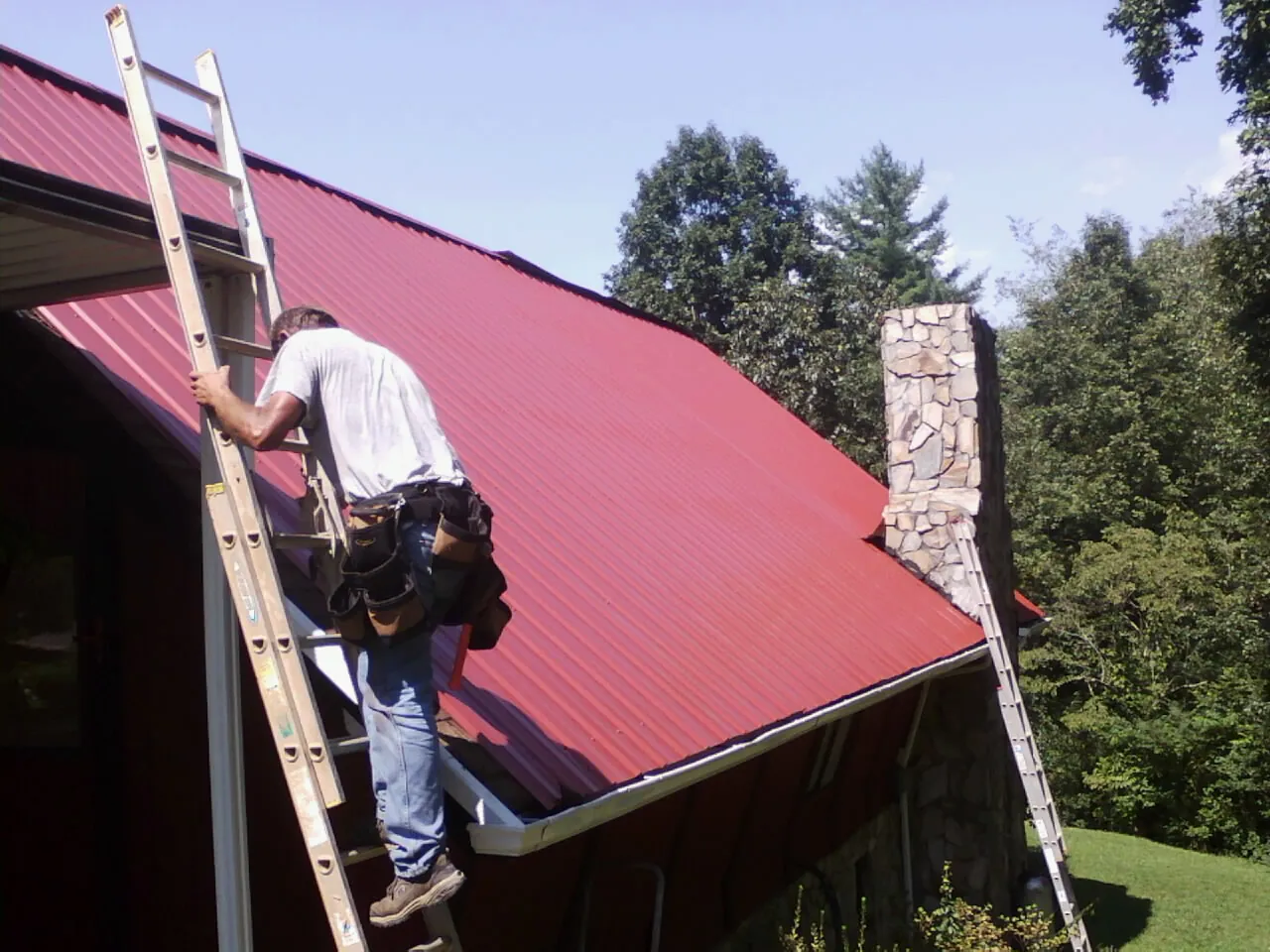 Roofer on a ladder beside a red metal roof and stone chimney — Soffit & Fascia Repair by Roofing Brothers in Fort Bridger